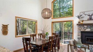 A dining area with a wooden table and chairs, large windows, and a stone fireplace. - The Retreat@ the Cottages-tucked in towering pines (McCall)