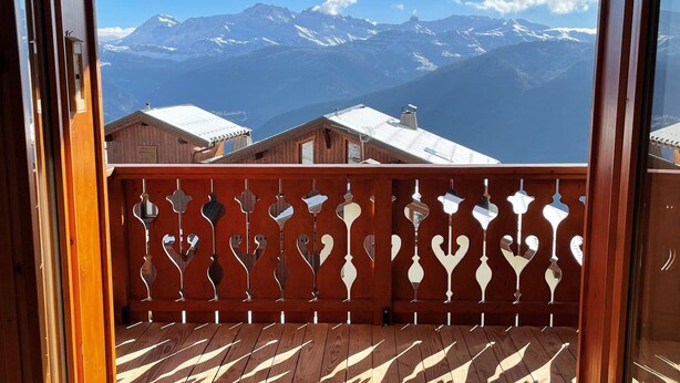 A wooden balcony with a patterned floor and a decorative railing, overlooking a mountainous landscape with snow-capped peaks.