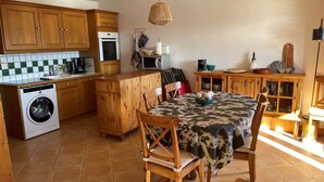 A compact kitchen with wooden cabinets, a washing machine, a dining table with chairs, and a hanging light fixture.