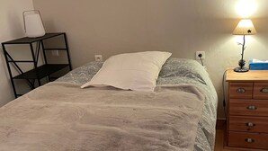 A neatly made bed with a white pillow and a gray blanket, a bedside table with a lamp, and a wooden dresser with drawers.