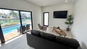 A modern living room with a black leather sofa, a wooden coffee table, and a TV mounted on the wall. A large window offers a view of a swimming pool and a building with palm trees.