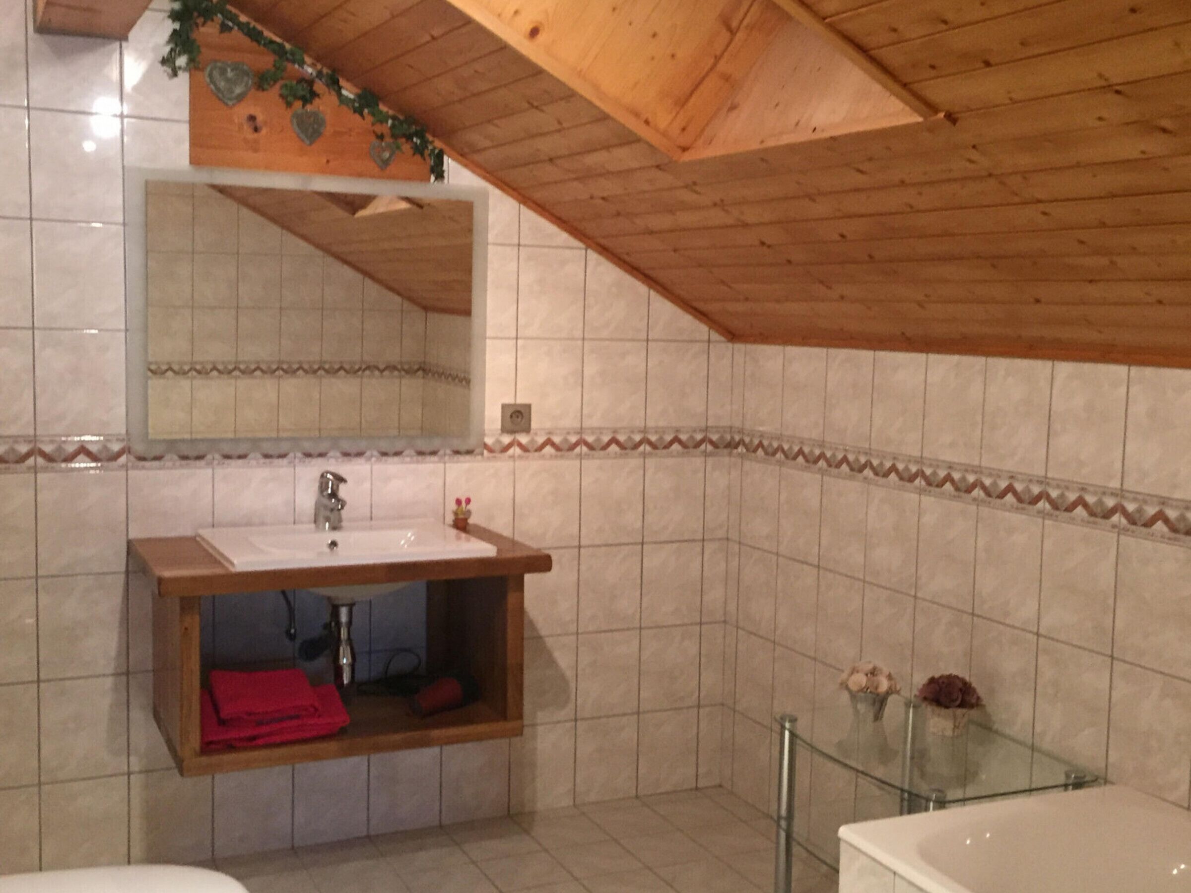 A bathroom with a wooden ceiling, a sink with a mirror, a glass shower enclosure, and tiled walls.