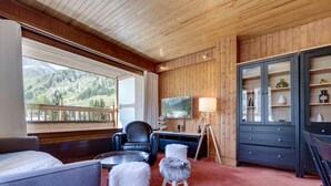 Une pièce chaleureuse avec un plafond en bois, une grande fenêtre donnant sur les montagnes, un bureau avec une lampe, une télévision et un meuble contenant des objets décoratifs.