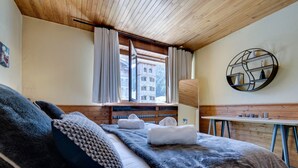 Une chambre d’hôtel avec un plafond en bois, un grand lit avec une couverture et des oreillers, une fenêtre avec des rideaux, un bureau en bois et une étagère fixée au mur, ornée d’objets décoratifs.