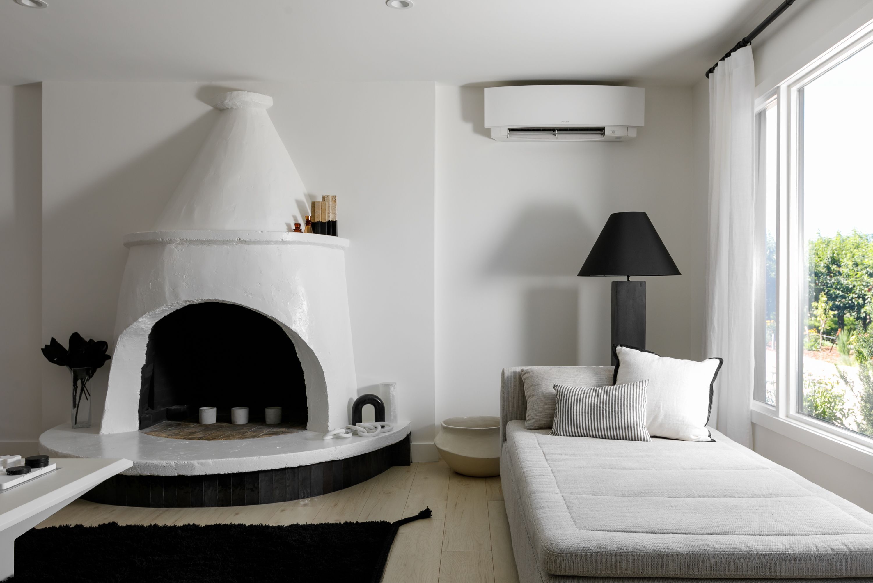 A modern living room with a fireplace, a sofa, a lamp, and a window with a view of greenery.