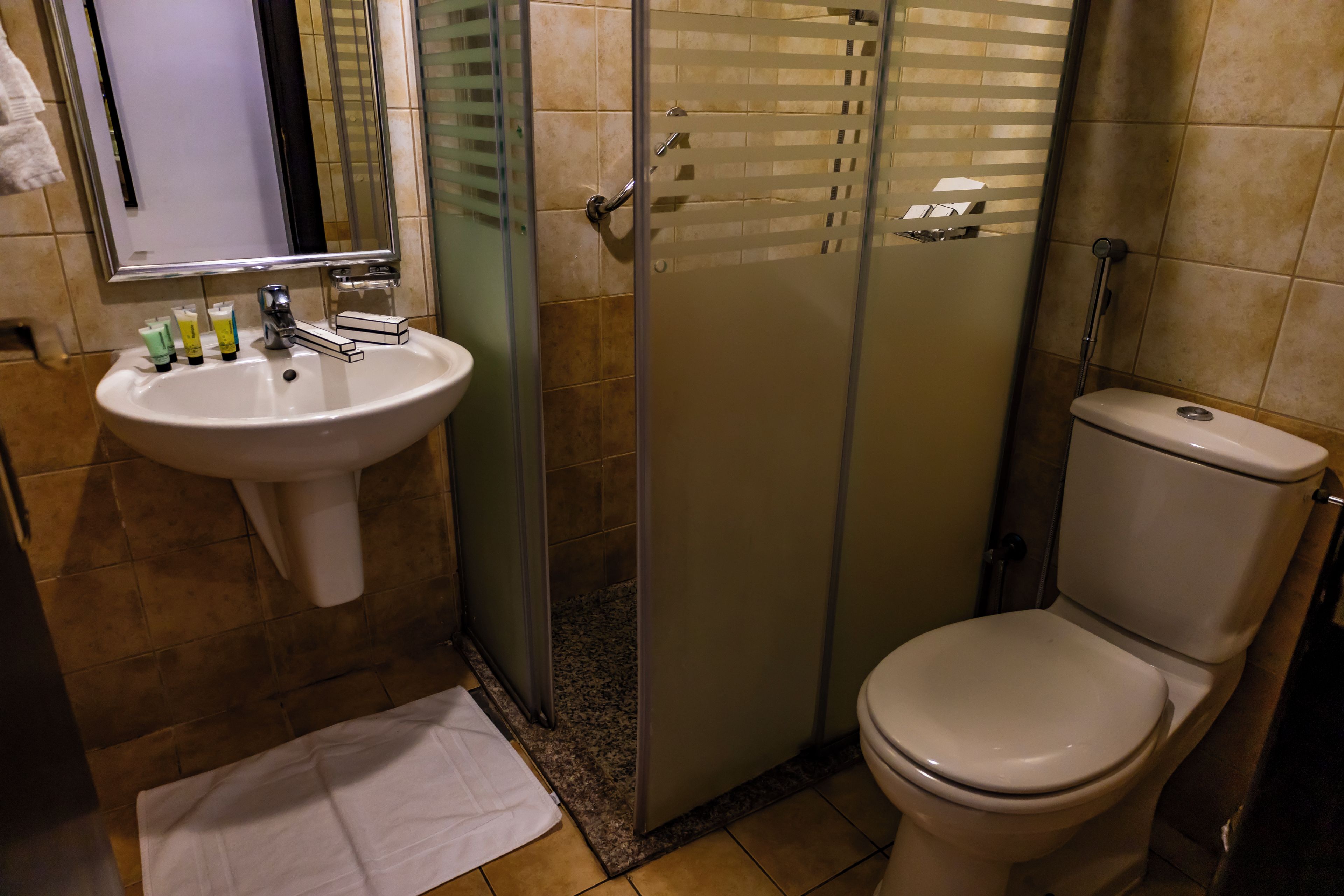 Un baño con lavamanos blanco, espejo e inodoro. Cuenta con un box de ducha de vidrio y un tapete de baño blanco en el suelo.