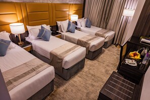 A hotel room with three beds, a nightstand, a desk with a lamp, and a small table with fruit and a bottle. - Al Mukhtara International Hotel (Madinah)