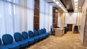 A conference room with rows of blue chairs, a carpeted floor, a projector screen, and a wooden desk.