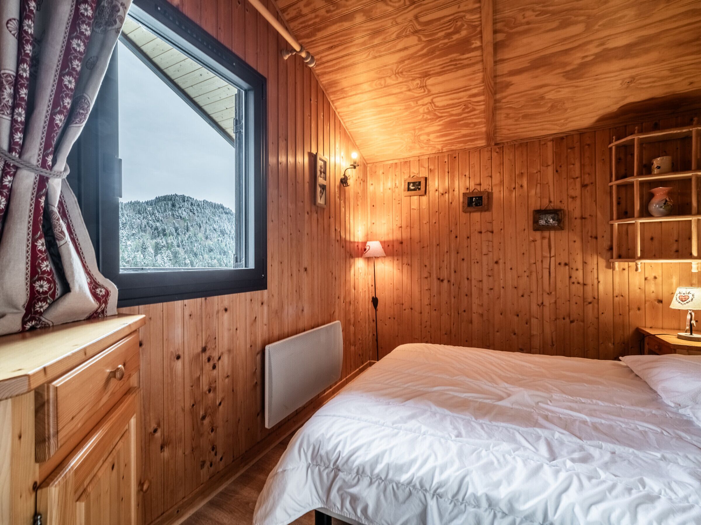 Une chambre dans un chalet en bois, avec un lit, une fenêtre avec des rideaux, une lampe de chevet et une étagère ornée d’objets décoratifs.