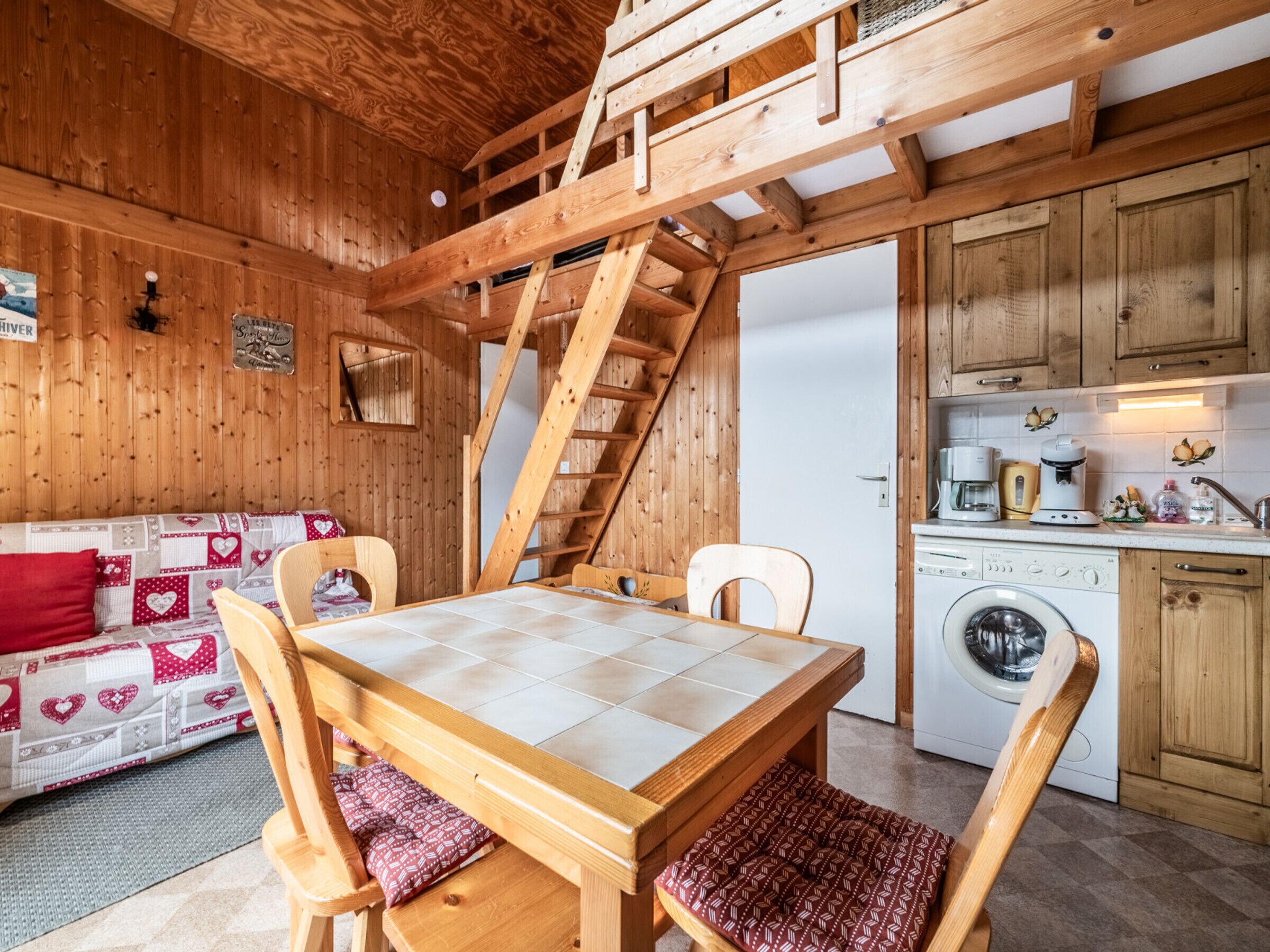 A cozy wooden cabin interior with a dining table, chairs, a sofa, a washing machine, and a kitchen area.