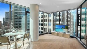 Ein modernes Wohnzimmer mit großem Fernseher, einem Essbereich mit Tisch und Stühlen sowie großen Fenstern mit Blick auf die Stadt.