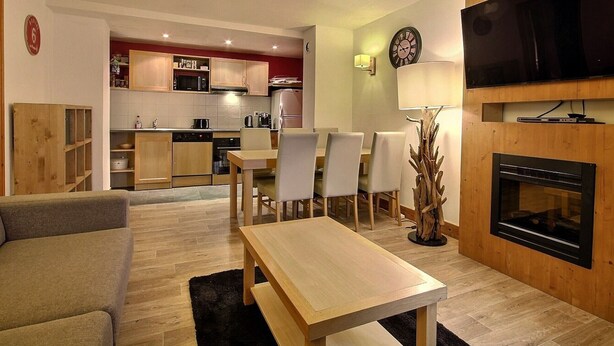 Una sala moderna con un sofá, una mesa de centro de madera y un comedor con sillas y una mesa. Se observa una cocina al fondo con armarios y electrodomésticos.