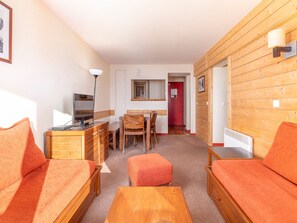 Un salon chaleureux avec un canapé, un pouf et une table basse en bois. On y trouve un téléviseur posé sur un meuble en bois, une lampe, ainsi qu’un coin repas avec une table et des chaises.