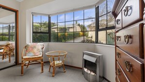 A room with a large bay window, wicker furniture, a fireplace, and a wooden dresser. - Central Comforts - Mount Eden Holiday Home (Auckland)