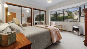 A bedroom with a bed, a chair, a table, a fireplace, and large windows. - Central Comforts - Mount Eden Holiday Home (Auckland)
