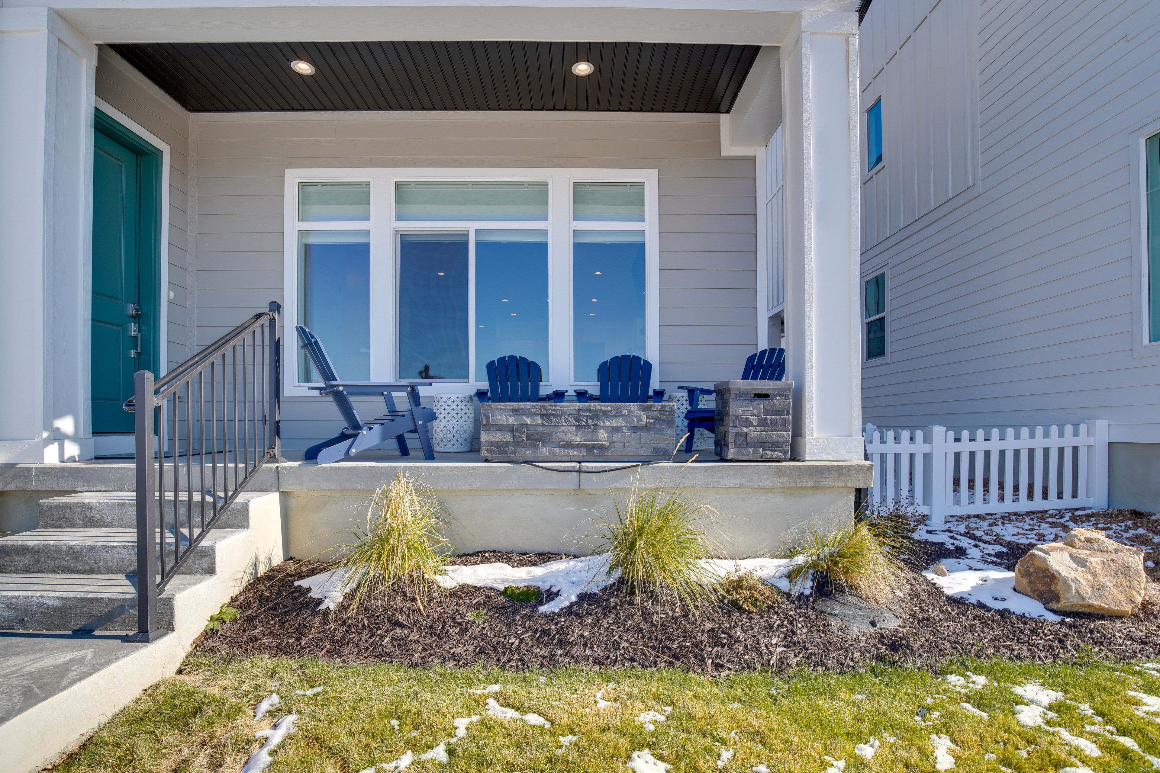 A covered porch with a stone bench and Adirondack chairs, a white picket fence, and a snow-covered lawn.
