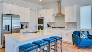 Una cocina moderna con armarios blancos, electrodomésticos de acero inoxidable, una isla central con taburetes y un sillón de color azul.