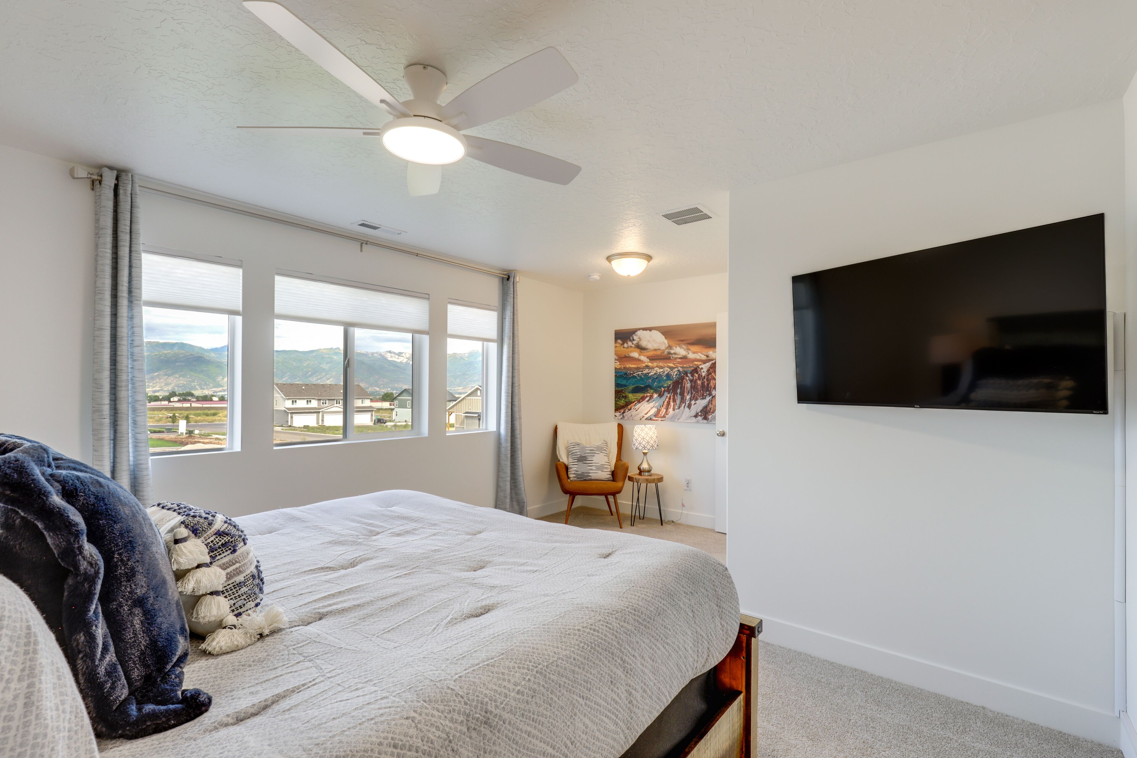 A bedroom with a large bed, a ceiling fan, a TV mounted on the wall, and a window with a view of the outdoors.