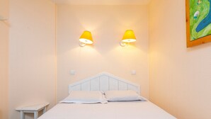 Un dormitorio con una cama blanca, dos lámparas de pared, una mesita de noche pequeña y un cuadro en la pared.