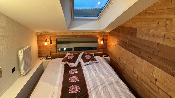 Ein Schlafzimmer mit schrägverdeckter Decke, Holzwänden, einem Oberlicht, einem Bett mit Zierteilen, Nachttischlampen und einer Heizung.