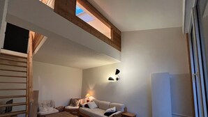Ein gemütliches Zimmer mit Schlafplatz in der Schlaffküche, einer Couch, einem Couchtisch und einem kleinen Tisch mit Lampe. Es gibt ein Fenster mit Vorhängen und einen am Wand montierten Fernseher.