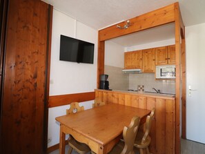 A compact kitchen with wooden cabinets, a flat-screen TV mounted above the counter, and a dining table with chairs.