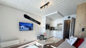 Un salon moderne avec un canapé, une table basse, un téléviseur fixé au mur et un coin cuisine visible à travers une porte ouverte.