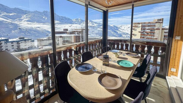 Un coin repas avec une table dressée pour quatre personnes, de grandes fenêtres offrant une vue sur les montagnes et un plafond en bois.