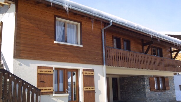 Una casa de madera de dos pisos con un balcón, base de piedra y techo cubierto de nieve.