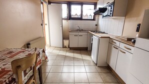 Una cocina compacta con armarios blancos, piso de baldosas, ventana con vistas y una mesa con mantel estampado.