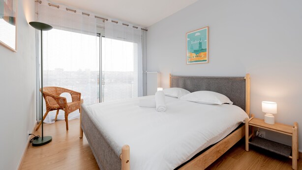 A modern hotel room with a large bed, a chair, a nightstand, and a city view.