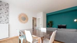 A modern living room with a teal accent wall, a wooden dining table with white chairs, a grey sofa, and a TV mounted on the wall.