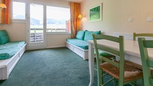 A room with a bed, a sofa, a dining table, and chairs. There is a window with curtains, a painting on the wall, and a view of the outdoors.