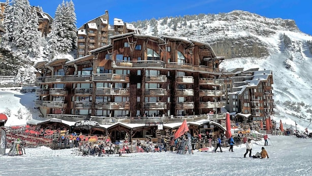 A multi-story wooden lodge with balconies, surrounded by snow-covered mountains and a ski slope.