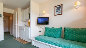 A room with a green sofa, a television mounted on the wall, and a kitchen area visible through an open door.