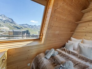 A wooden cabin with a bed, pillows, and a window offering a mountain view.