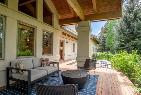 An outdoor deck with a sofa, chairs, and a table, surrounded by greenery and a building with large windows.