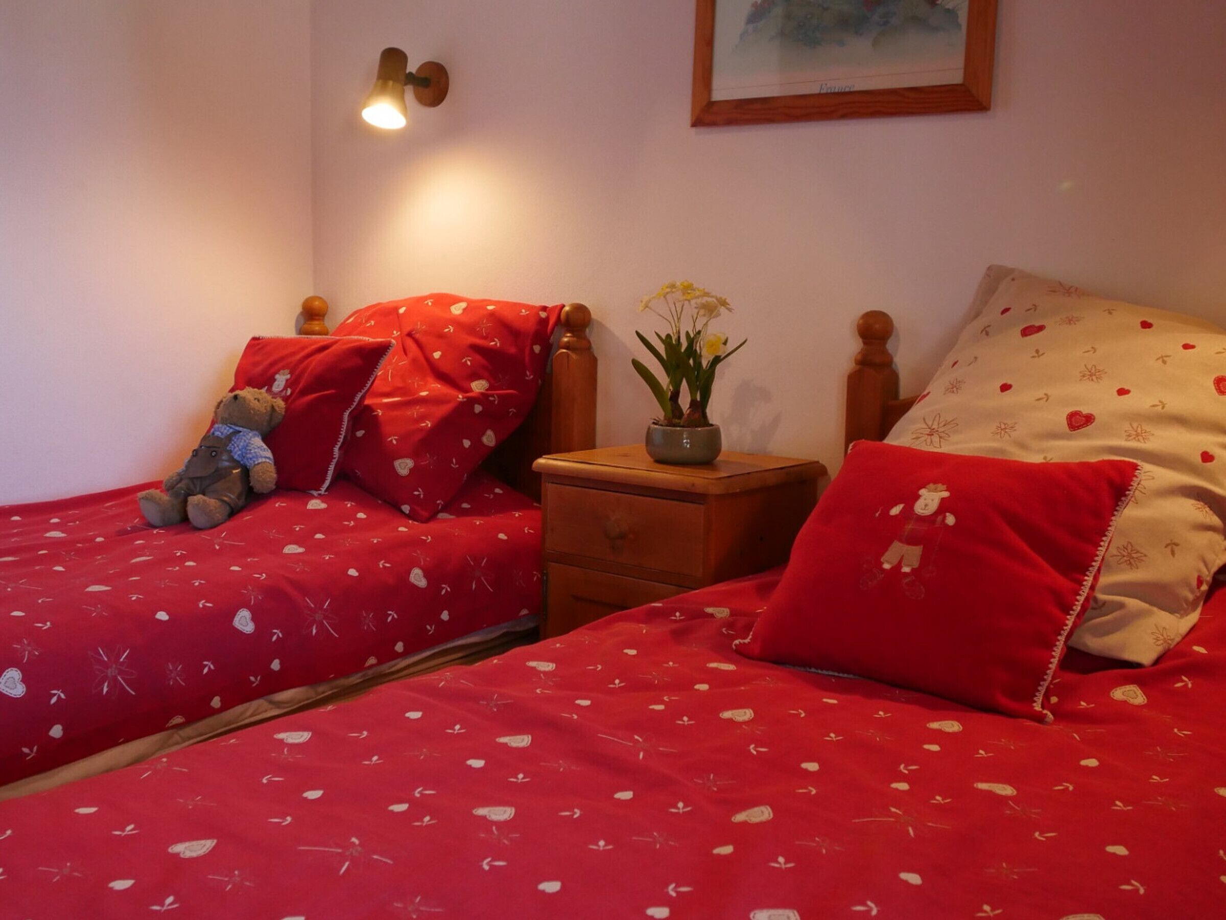 A bedroom with two beds, each with red bedding and heart patterns, a teddy bear on one bed, a nightstand with a lamp, and a framed picture on the wall.