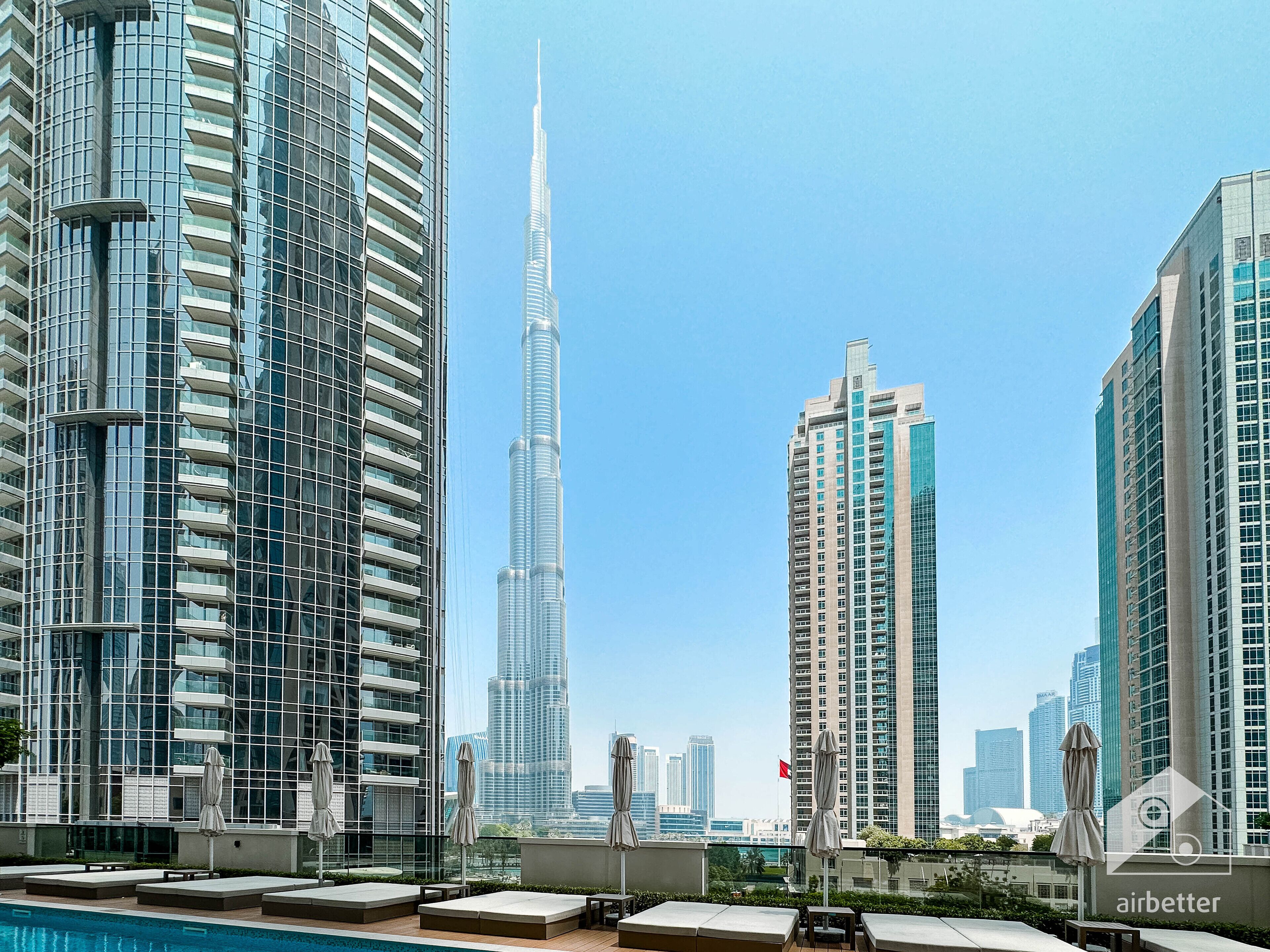 A rooftop pool area with lounge chairs and umbrellas, overlooking modern high-rise buildings and a prominent skyscraper.