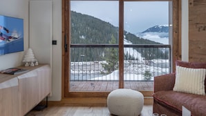 Ein Zimmer mit Holzboden, einem weißen Hocker, einem braunen Sofa mit weißem Kissen, einem an der Wand befestigten Fernseher und einer großen Schiebeglasdoor mit Blick auf die Berge.