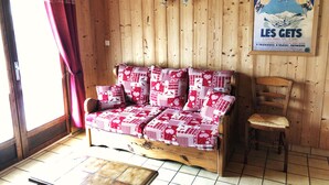 Interior de una cabaña de madera con sofá y sillón, un cartel en la pared y una ventana con cortinas.