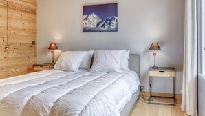 A bedroom with a bed, two bedside tables, a wall-mounted lamp, and a painting of a mountain.