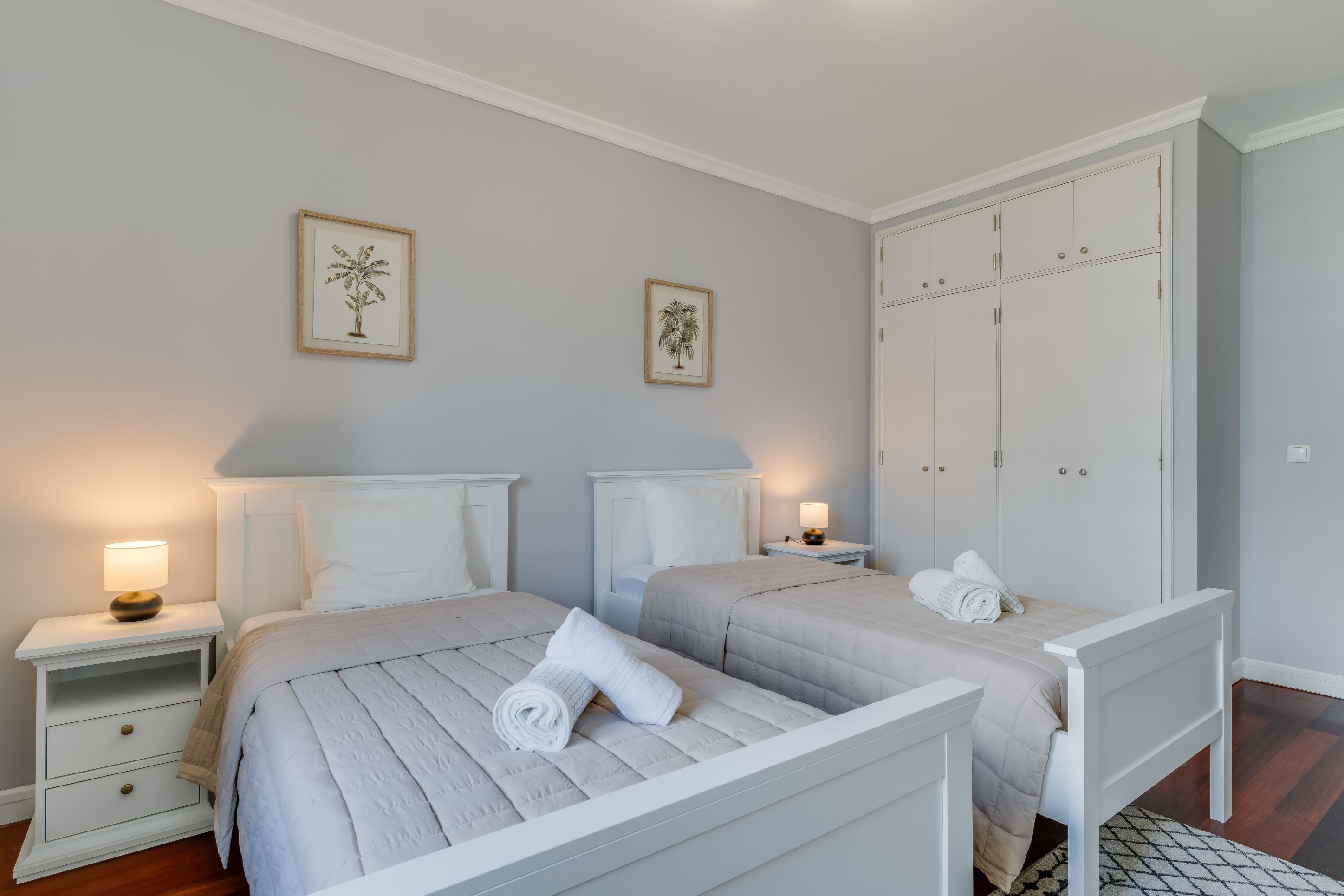 A bedroom with two beds, white nightstands, a wardrobe, and framed pictures on the wall.