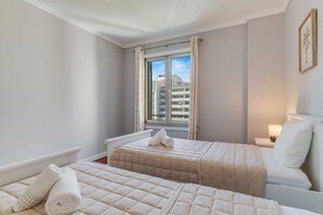 Hotellihuone, jossa on kaksi sänkyä, ikkuna, jossa on verhot, yöpöytä lampun kanssa ja seinällä oleva kehystetty kuva.