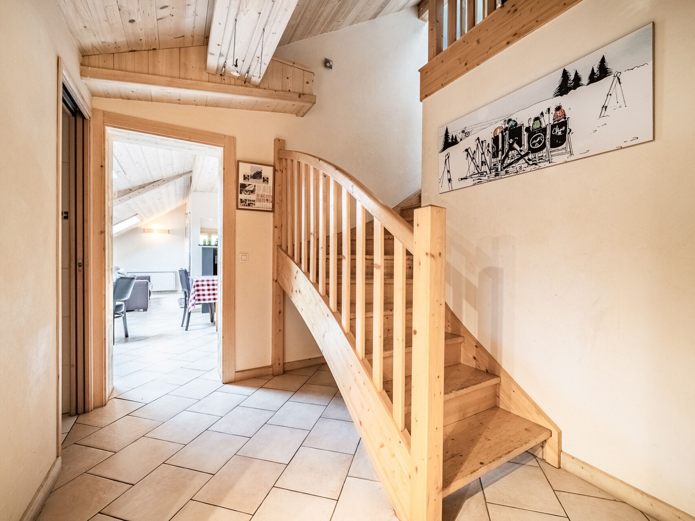 Eine Holztreppe führt zu einer zweiten Etage, ein Essbereich mit kariertem Tischlein und ein an der Wand befestigtes Schild mit einer Darstellung von Skifahrern.