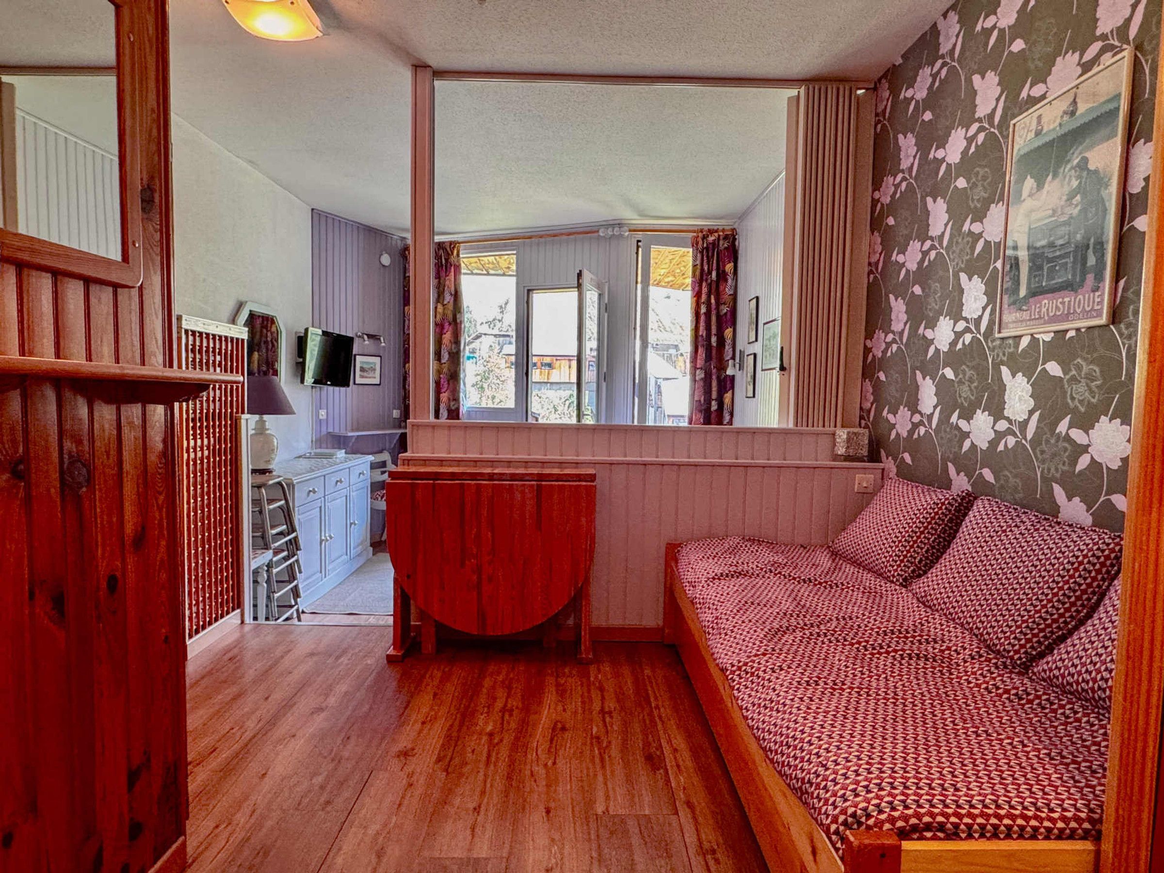 A cozy room with a wooden floor, a red bench, a patterned sofa, and a view of the outside through a window.
