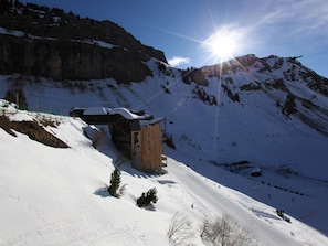 A wooden cabin on a snowy mountain slope with a clear sky and a bright sun.