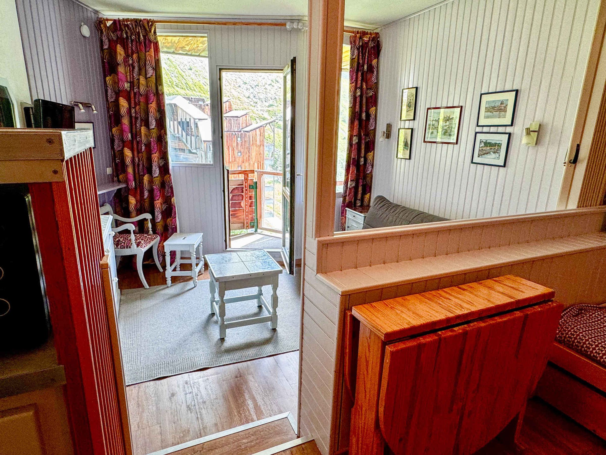 A room with a wooden bench, a small table, and a chair. There is a door leading to an outdoor area with a view of buildings.