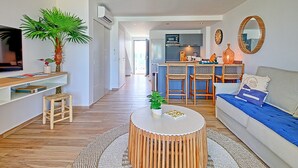 A modern living room with a TV, a large plant, a round coffee table, and a kitchen in the background.