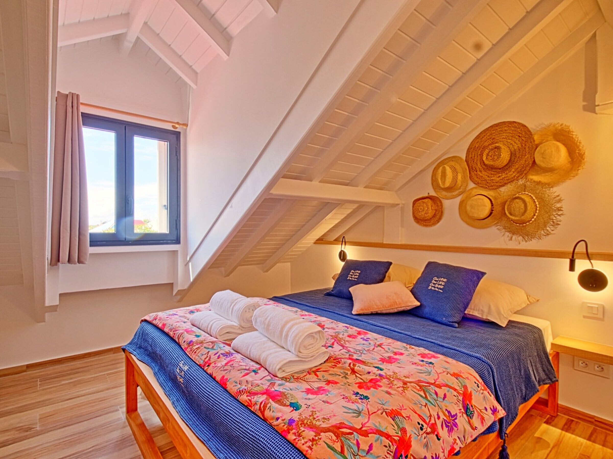 Une chambre avec un plafond en pente, un lit recouvert d’une couvre-lit à motifs floraux et une fenêtre avec des rideaux.